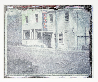 A white clapboard covered building with a blue and red sign that reads Hotel. There is a car parked on the street. Overall the image is grayish and distressed.