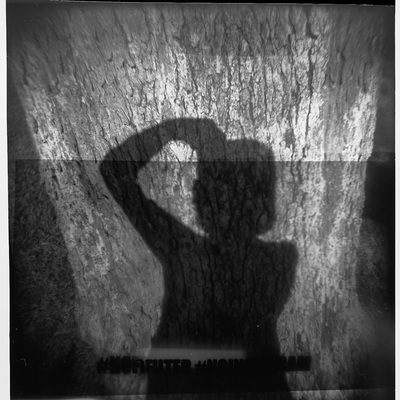 Black and white image of photographer's shadow against a tree. Blurry black text at the bottom reads `#nofilter #noinstagram`