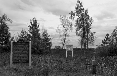 A black and white photo with a historical marker for Alabaster. Next to it is a worn 'Keep Out' sign. There is a fence in the foreground, and forbidding clouds in the background.