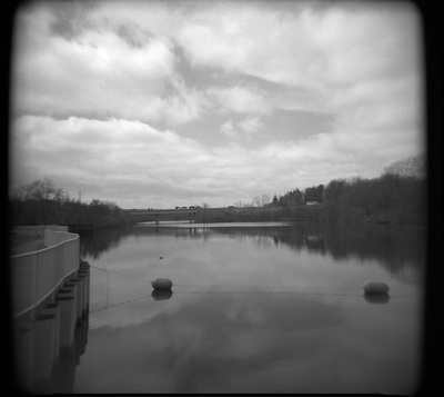 Square image of smooth water with two buoys in it; dramatic clouds overhead. The edges of the frame are dark.