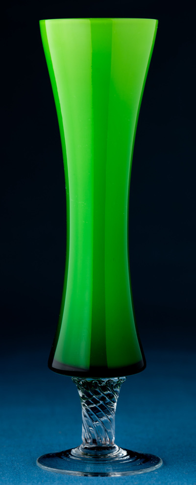 A slender lime green vase with clear footed base stands on a blue table with a dark background. It is flared on the top and curves in at the middle.