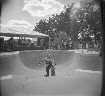 A woman with braided hair is alone in the bowl, her skateboard visible over the edge. A band plays under a canopy.