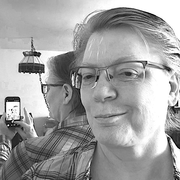 A person wearing glasses and a plaid shirt takes a selfie with a smartphone, showing their reflection in a mirror. The reflection captures the person holding the phone.