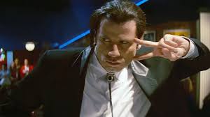 John Travolta holding two fingers in a v-shape pointing at the side of his head